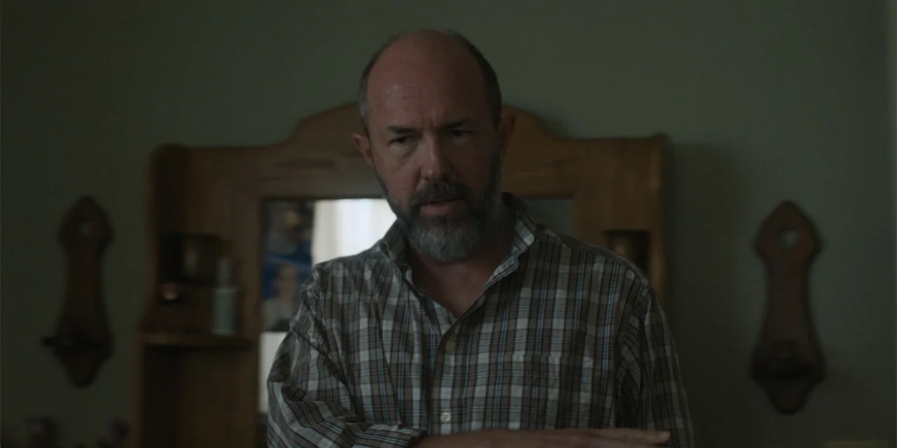 Caddo Lake (2024) movie still: medium — A dimly lit residential bedroom features a wooden dresser with an attached mirror. A middle-aged man with a…