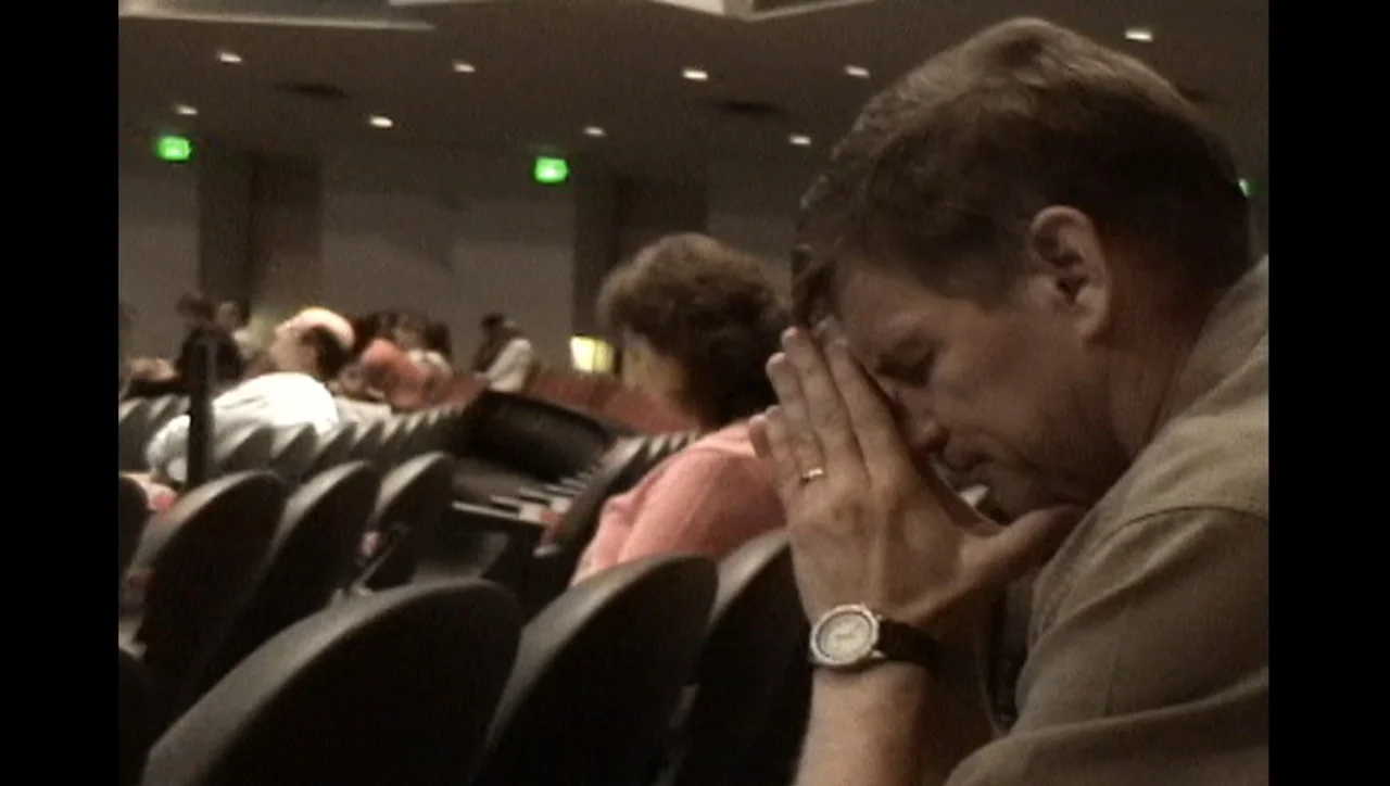 The Trials of Ted Haggard (2009) movie still: medium — A dimly lit auditorium contains rows of dark chairs and a seated audience. A man sits in the…
