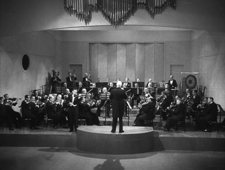 To Joy (1950) movie still: wide — An indoor concert stage features a large orchestra seated in rows before a pipe organ. The conductor directs the…