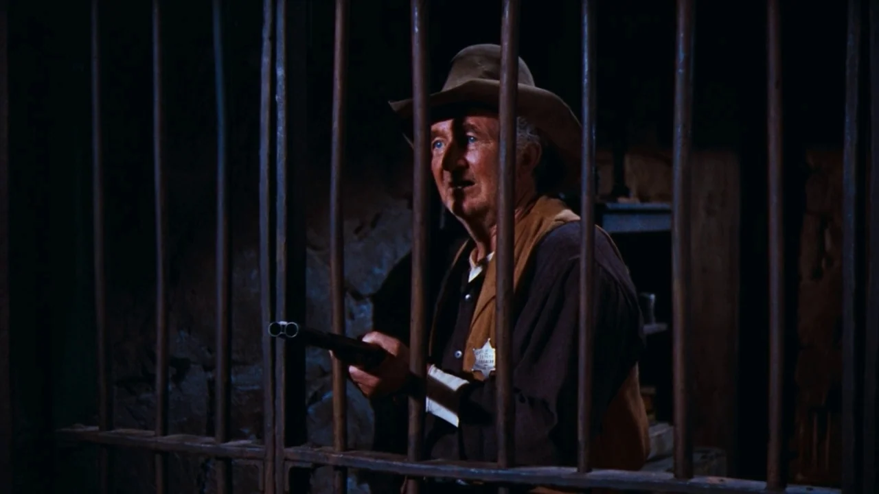 Rio Bravo (1959) movie still: medium — A dimly lit room features a person framed behind heavy vertical metal bars. An elderly man in a cowboy hat…