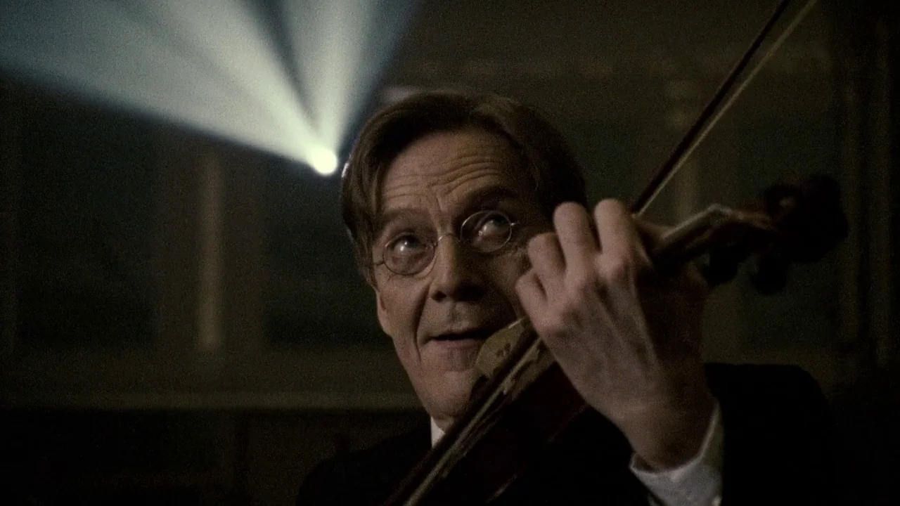 Everlasting Moments (2008) movie still: closeup — A man plays a violin in a dark, dimly lit indoor space. A single bright beam of light cuts through…