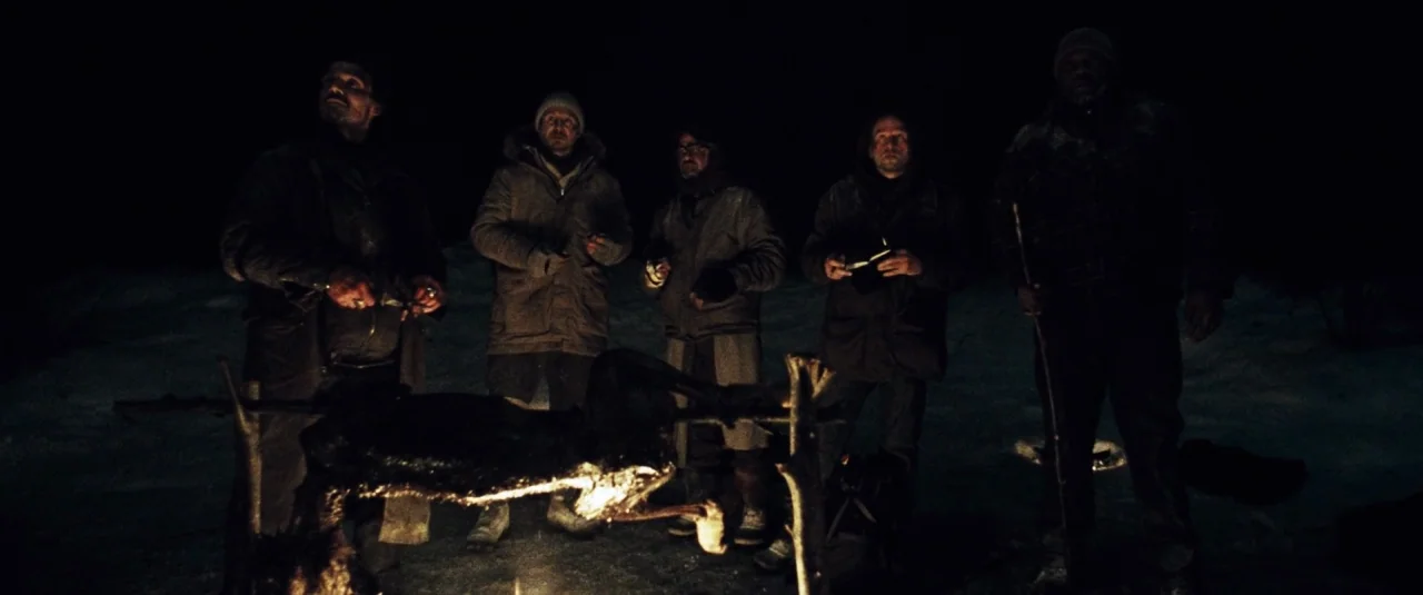 The Grey (2012) movie still: fullbody — A group of five men stands in a dark, snow-covered landscape at night. They gather around a small fire pit…