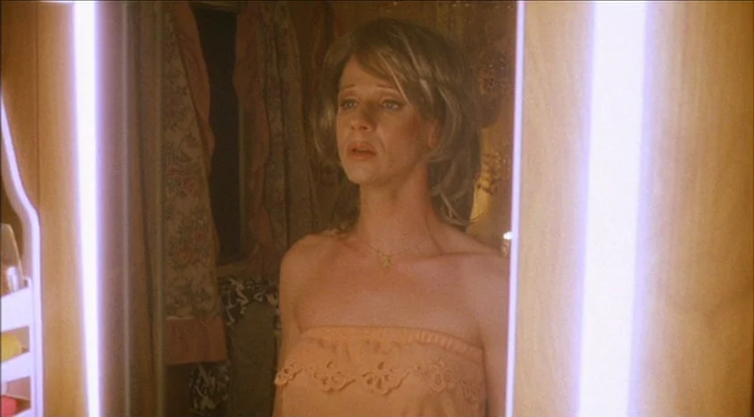 Hedwig and the Angry Inch (2001) movie still: medium — A person stands before a vertical vanity mirror in a dimly lit room. The subject wears a wig…