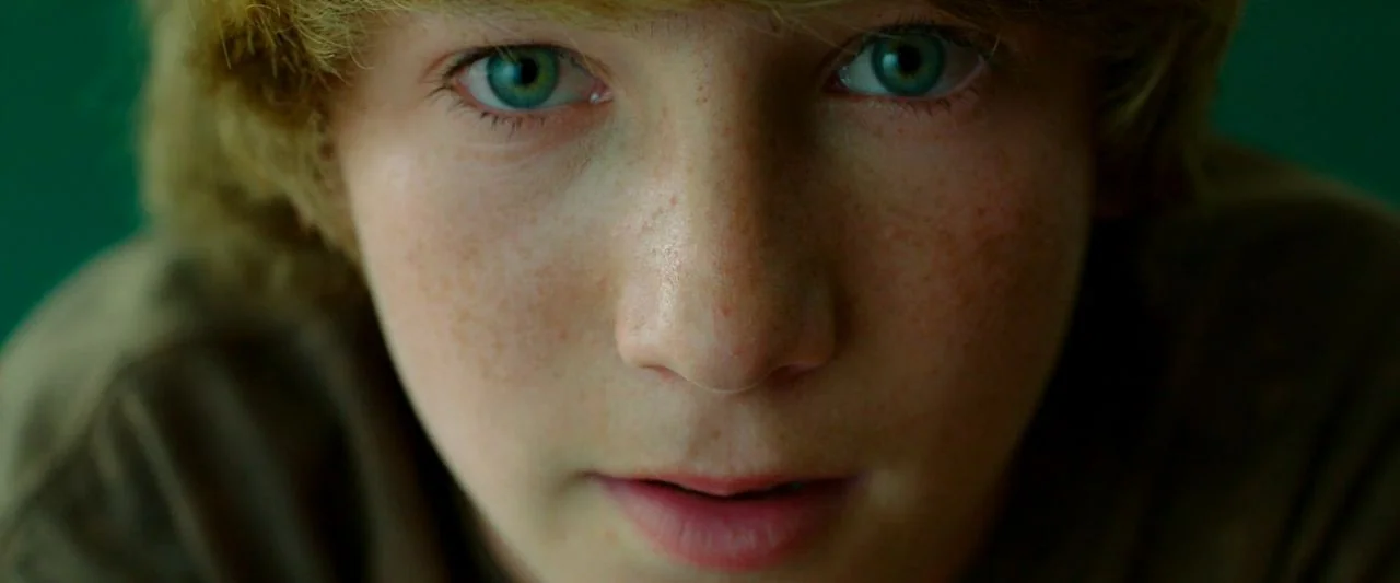 Cooties (2014) movie still: closeup — A close-up portrait of a young person against a plain, dark green wall. The subject faces the camera directly…