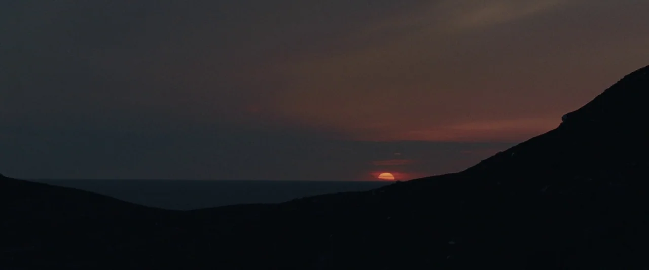 God’s Creatures (2022) movie still: establishing — The sun sets behind a dark silhouette of rolling hills overlooking the open ocean. A thin sliver…