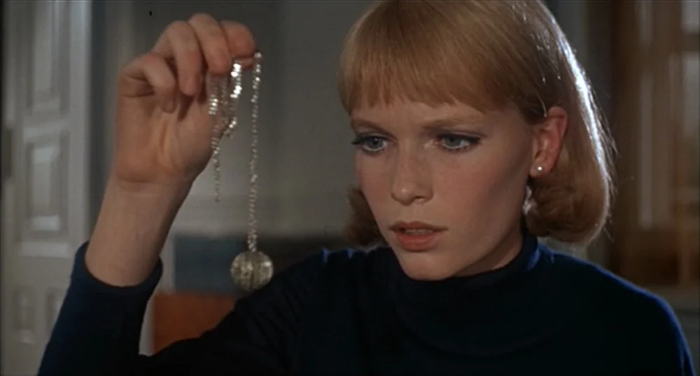 Rosemary’s Baby (1968) movie still: closeup — A woman sits in a room while examining a small object held in her hand. The background is slightly out…