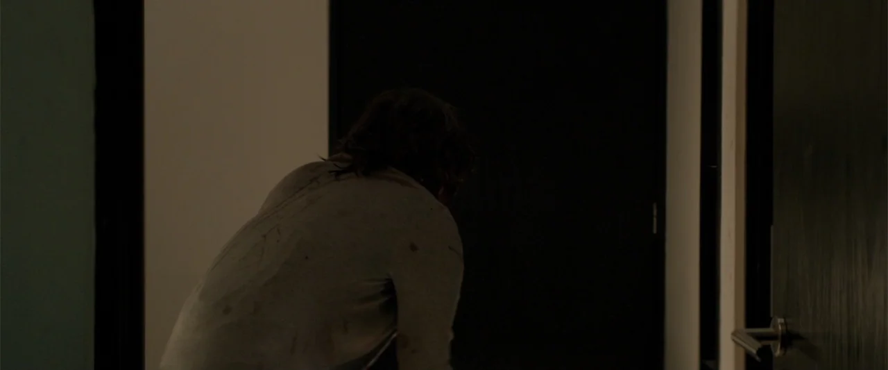 Human Animals (2020) movie still: medium — A dark, dimly lit hallway with white walls and a solid black closed door. A person wearing a stained grey…