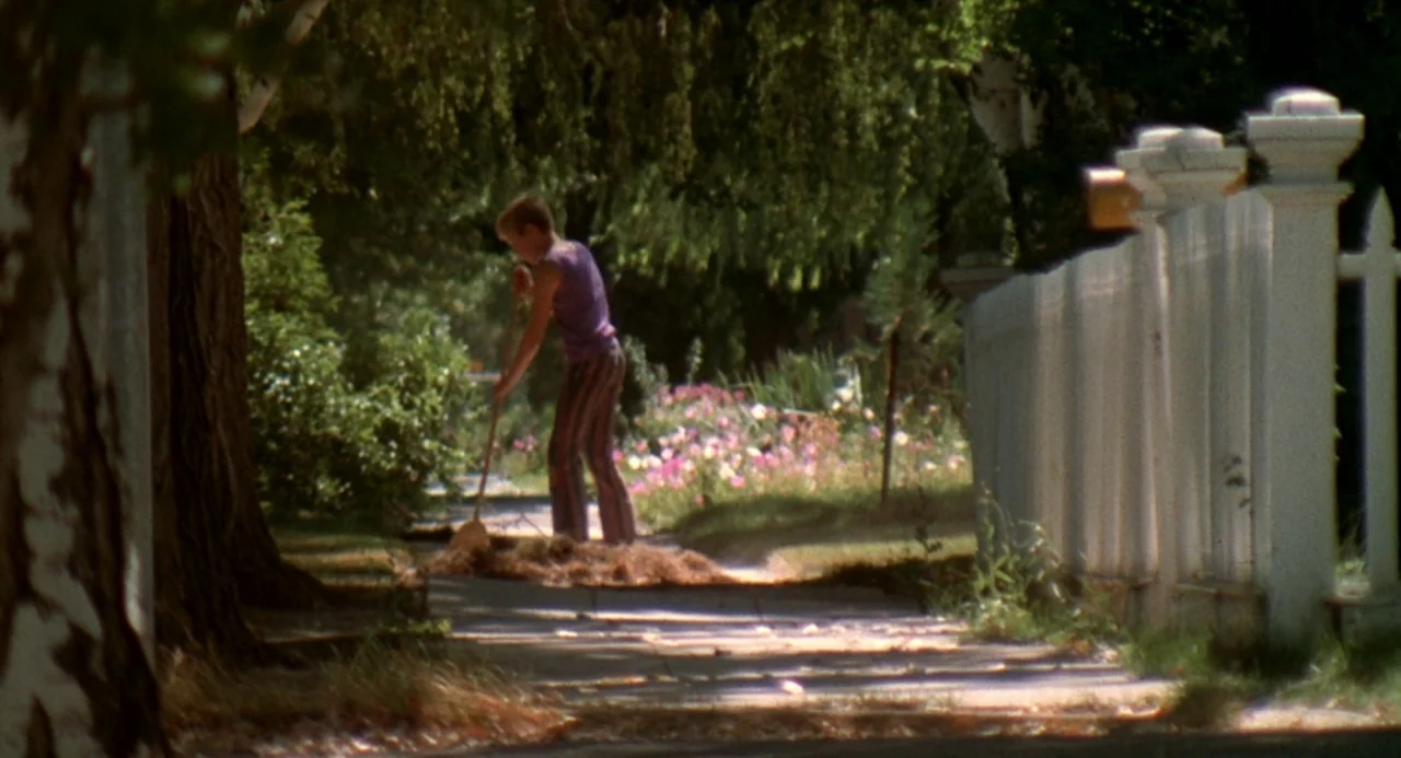 Charley Varrick (1973) movie still: wide — A residential sidewalk runs beside a white picket fence and a flower bed. A person uses a broom to sweep…