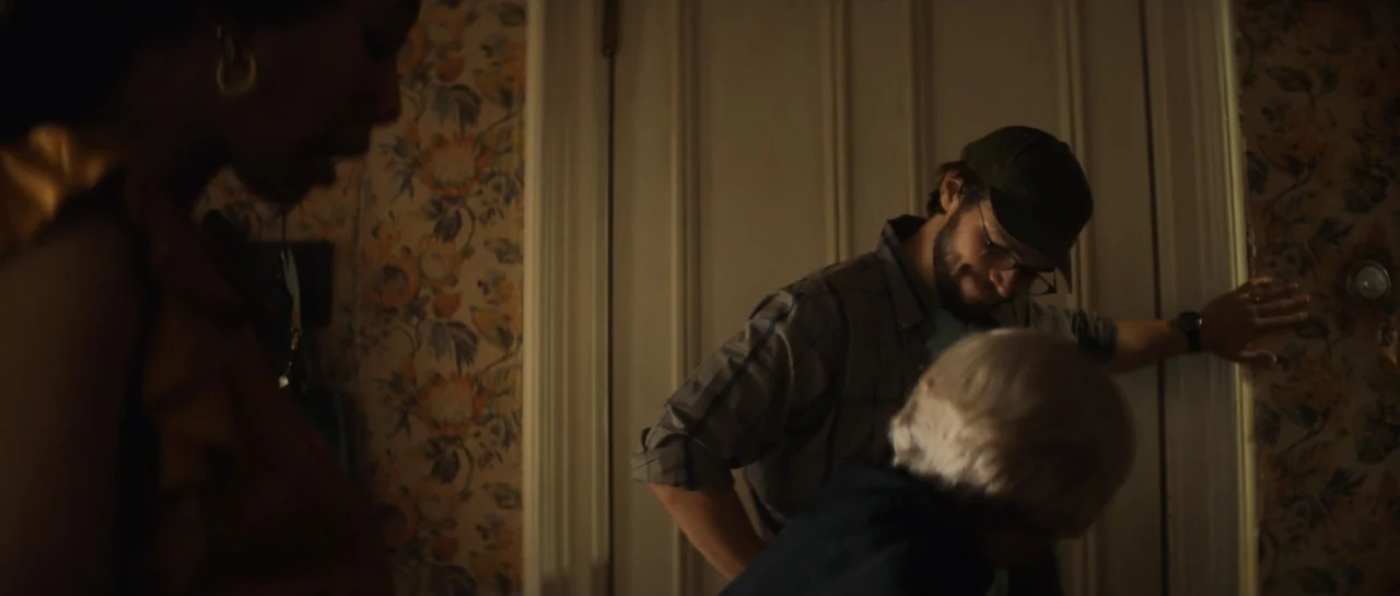 The Front Room (2024) movie still: medium — A dimly lit residential hallway with floral patterned wallpaper and a white wooden door. A man leans…