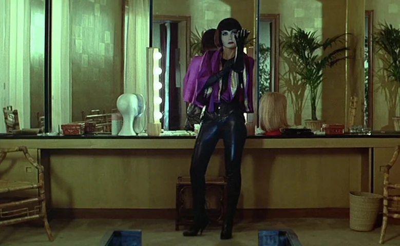 Maîtresse (1975) movie still: fullbody — A dressing room features a long wall mirror and a wooden vanity bench. A woman stands in front of the…