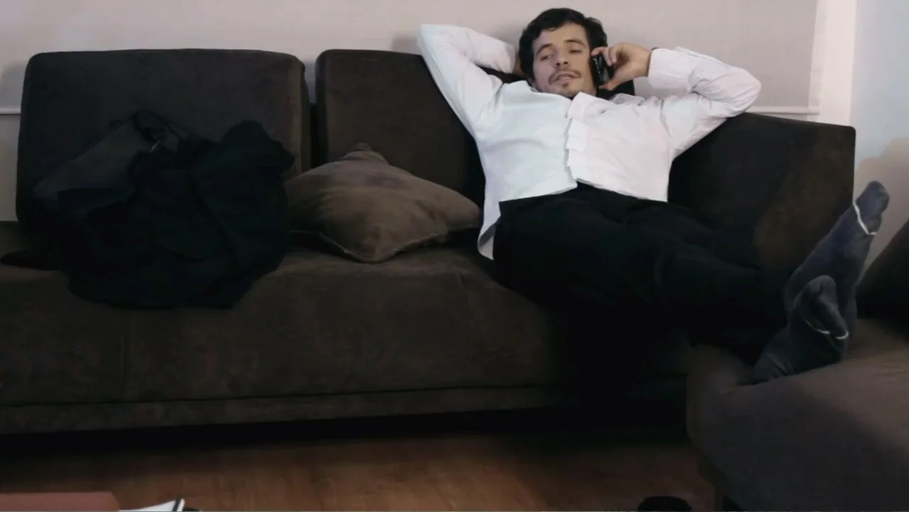 Aquel Septiembre (2018) movie still: medium — A living room with a brown sofa and a dark cushion. A man in a white shirt and black trousers lies on…