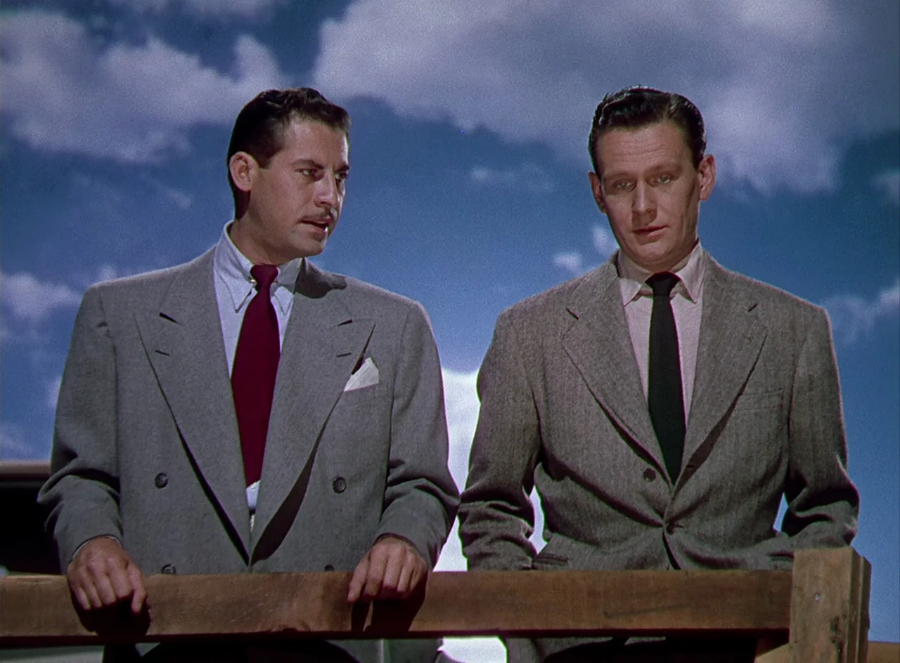 Desert Fury (1947) movie still: medium — Two men in business suits stand outdoors before a blue, cloud-filled sky. They lean against a wooden…