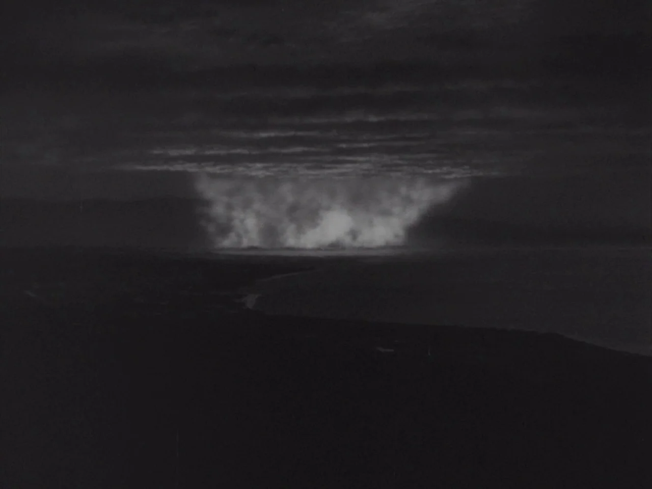 Godzilla Raids Again (1955) movie still: establishing — A dark coastline at night sits beneath a heavy, cloudy sky. A brilliant white light radiates…