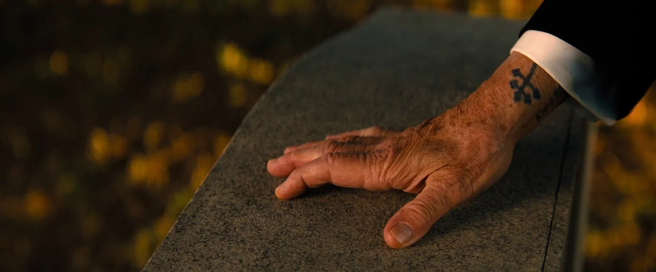John Wick: Chapter 4 (2023) movie still: closeup — A textured grey stone slab sits in a park during the late afternoon. An aged hand with a…