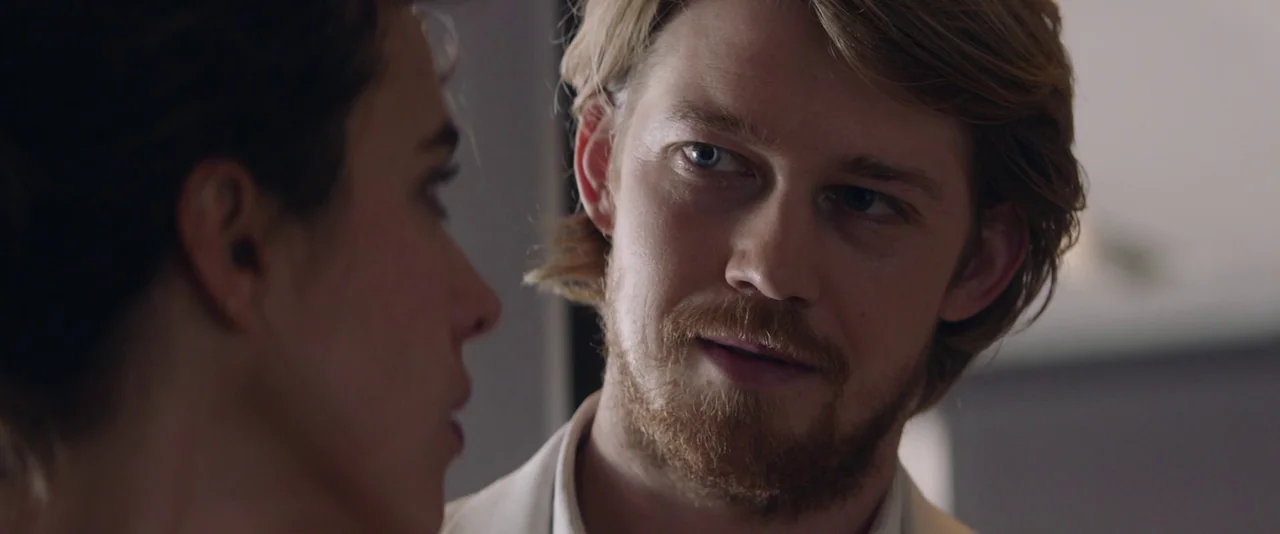 Stars At Noon (2022) movie still: closeup — An interior space with a man in a white shirt interacting with a woman. The man has blonde hair and a…