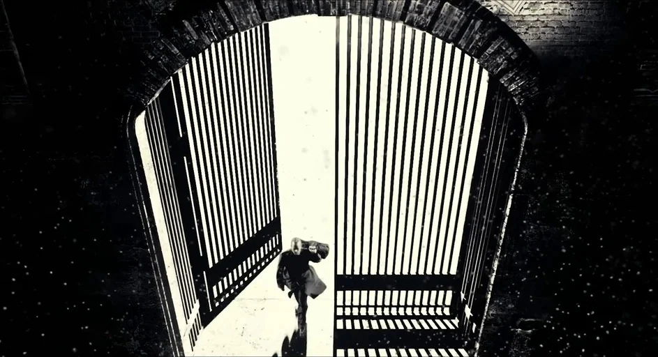 Sin City (2005) movie still: wide — A man walks through a large open metal gate at night. Floating particles are visible against the extreme bright…