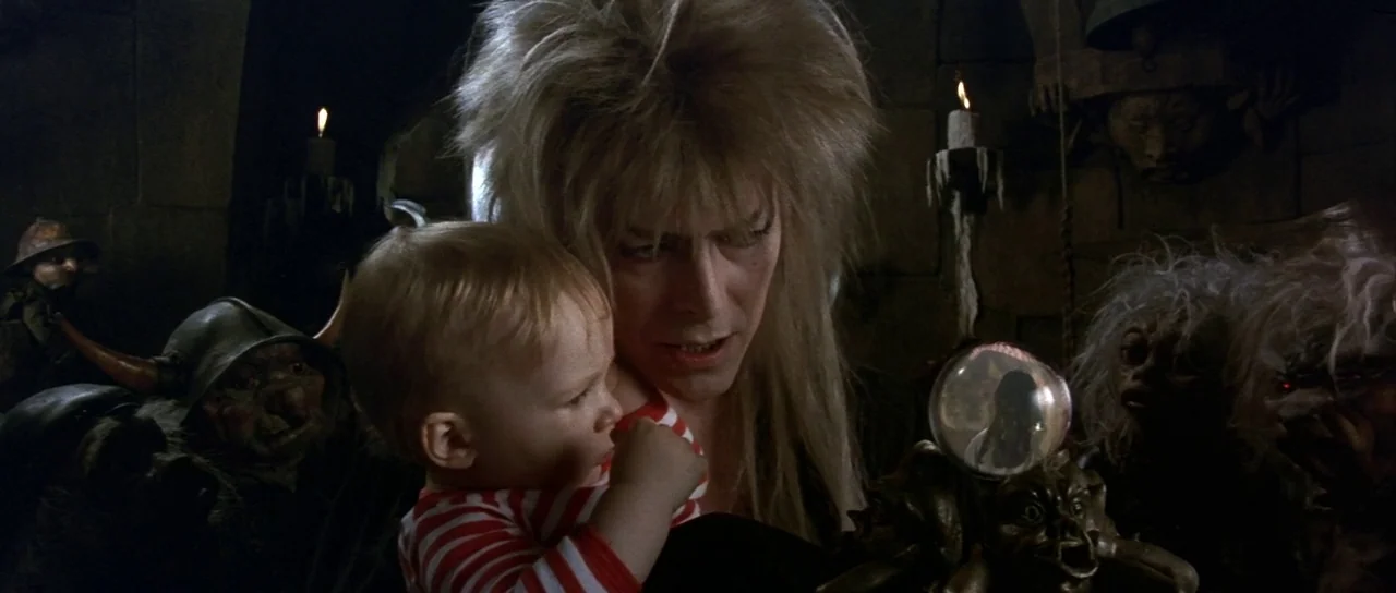 Labyrinth (1986) movie still: medium — A dark stone chamber lit by small flickering candles perched on nearby wall ledges. A man with elaborate…