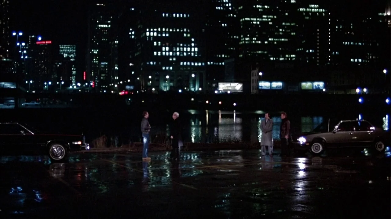 Thief (1981) movie still: wide — A wet asphalt parking lot at night bordered by a dark river and cityscape. Four men stand in pairs near their…