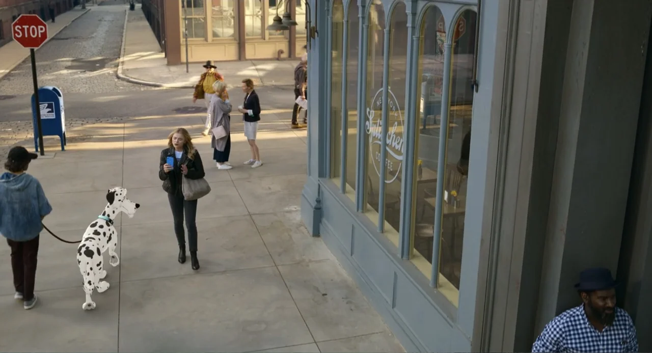 Tom & Jerry (2021) movie still: wide — A sunlit city sidewalk runs alongside a storefront with large, glass-paned windows. A woman walks toward the…