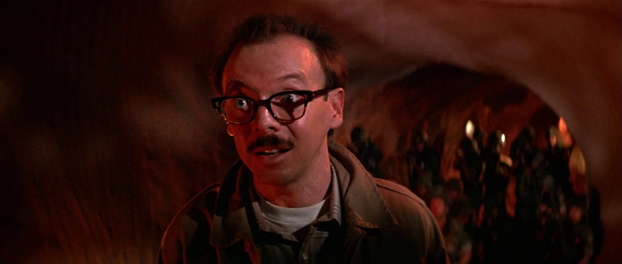 Invaders From Mars (1986) movie still: medium — A man stands inside a dimly lit rocky tunnel with organic, cavernous walls. He gazes off-camera with…