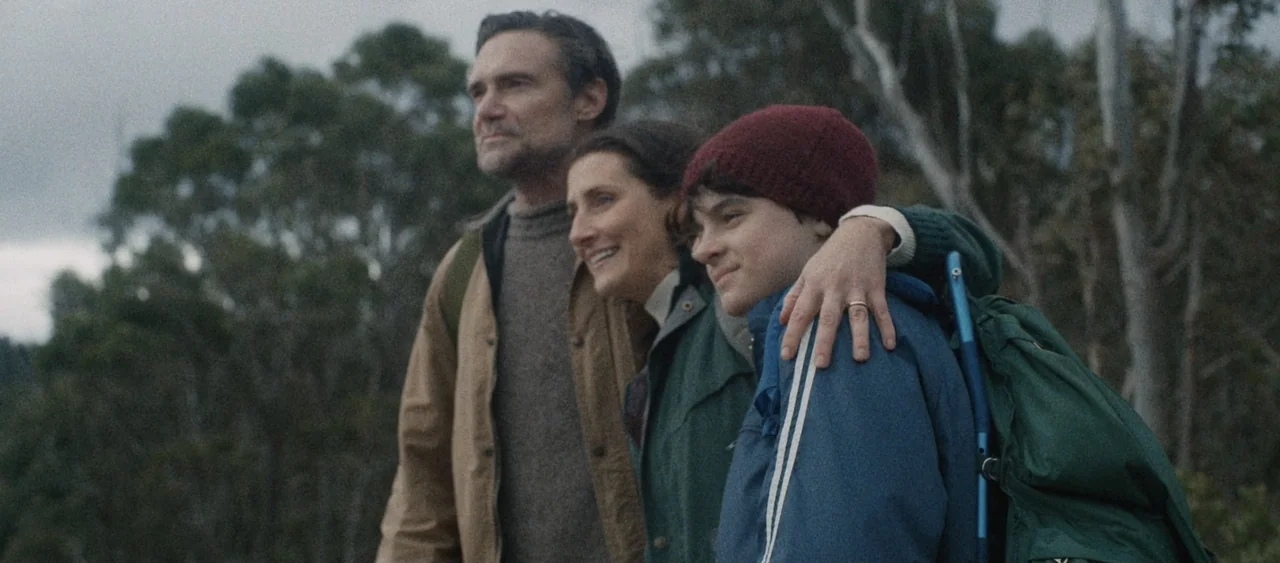 Force of Nature: The Dry 2 (2024) movie still: medium — A hillside landscape filled with tall trees stands behind three people standing together. A…