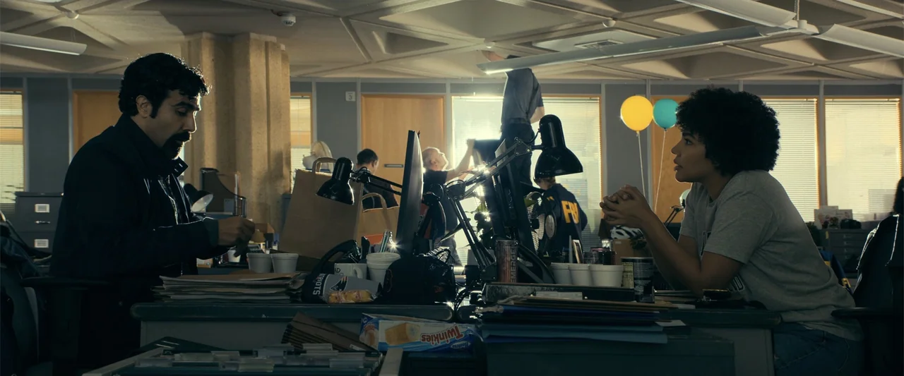 The Beekeeper (2024) movie still: medium — A cluttered office space features desks piled with files and office supplies in the foreground. A man and…