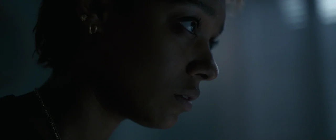 Talk to Me (2023) movie still: closeup — A dimly lit room with blue-tinted shadows and a bright off-screen light source. A young woman looks toward…