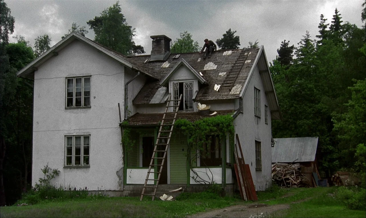 My Life As A Dog (1985) movie still: wide — A rustic two-story house with white stucco walls stands under a heavy overcast sky. A man sits atop the…