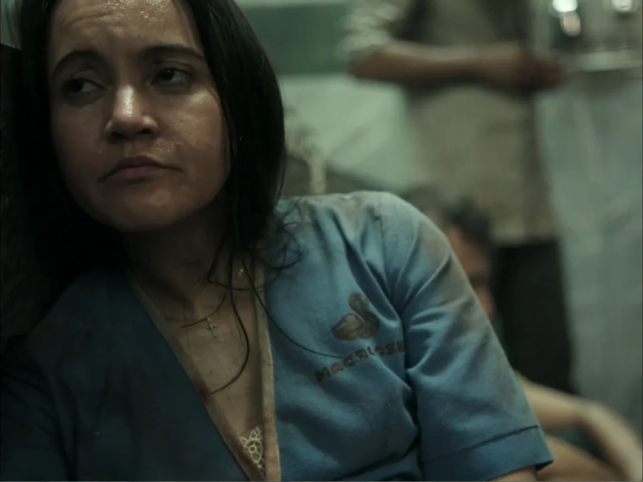 November (2025) movie still: medium — A dimly lit industrial room with blurred figures visible in the background. A woman in a blue uniform leans…