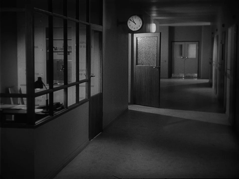 Brink of Life (1958) movie still: establishing — A dimly lit hospital corridor with glass-partitioned rooms and a wall-mounted analog clock. A…