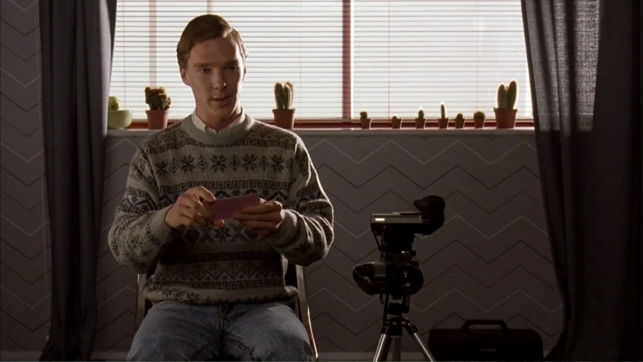 Starter for 10 (2006) movie still: medium — A man sits in a chair in front of a window with blinds. He holds a small pink paper card while a video…