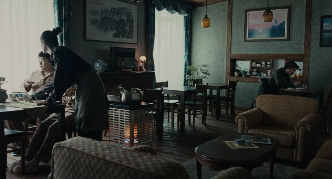 Winter in Sokcho (2025) movie still: wide — A cafe interior with wooden floors, patterned wallpaper, and several dining tables. People sit at tables…