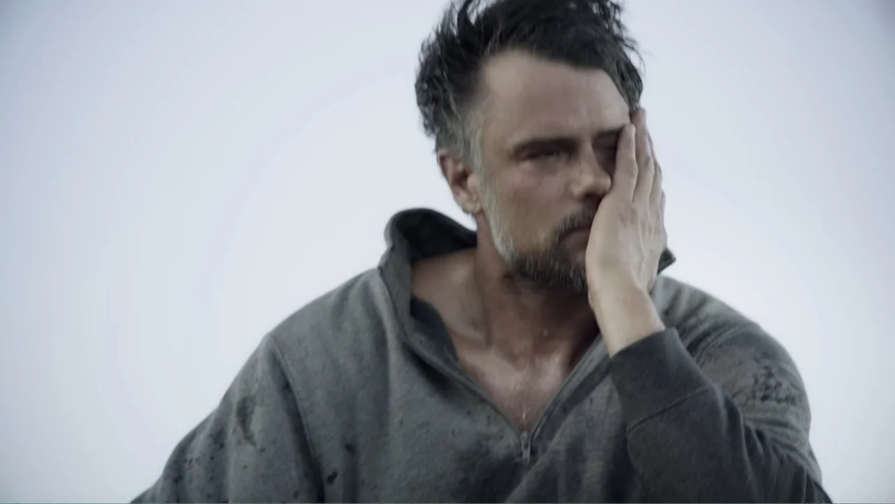 Capsized: Blood in the Water (2019) movie still: closeup — An outdoor area with a featureless, light-grey sky backdrop. A man in a dirty grey hoodie…