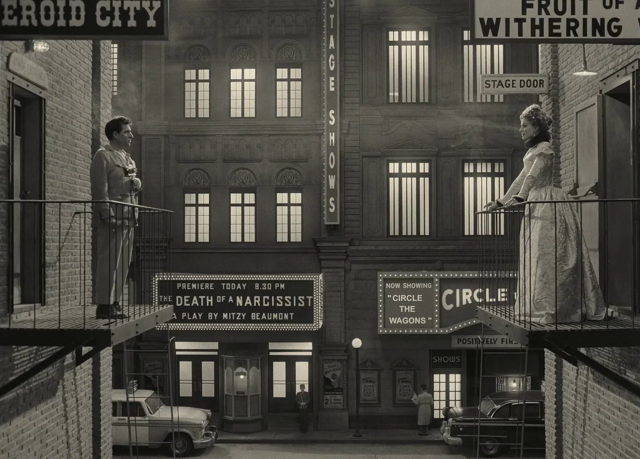 Asteroid City (2023) movie still: wide — A 1950s city street is flanked by brick buildings with fire escapes and signs. A man and a woman stand on…
