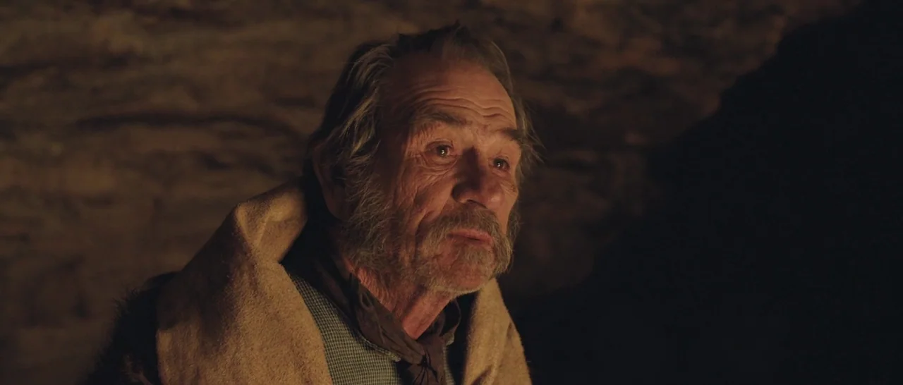 The Homesman (2014) movie still: closeup — A dark cave interior with rough, uneven rocky walls surrounding the subject. An elderly man is draped in…