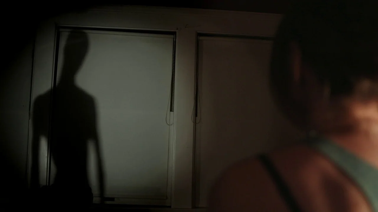 The Pact II movie still: medium — A dim room features two large window blinds covering a wall surface. A sharp, dark, elongated human silhouette is…
