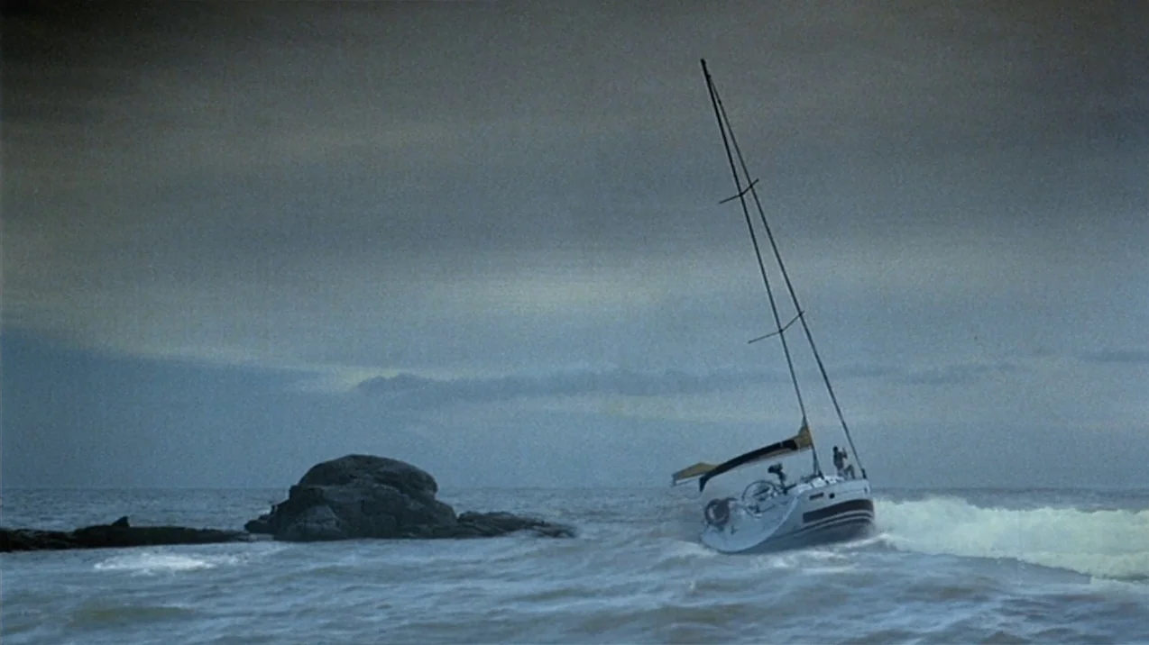Dagon (2001) movie still: wide — A sailboat lists sharply in turbulent water beside a large dark rock formation. Heavy grey clouds cover the sky…