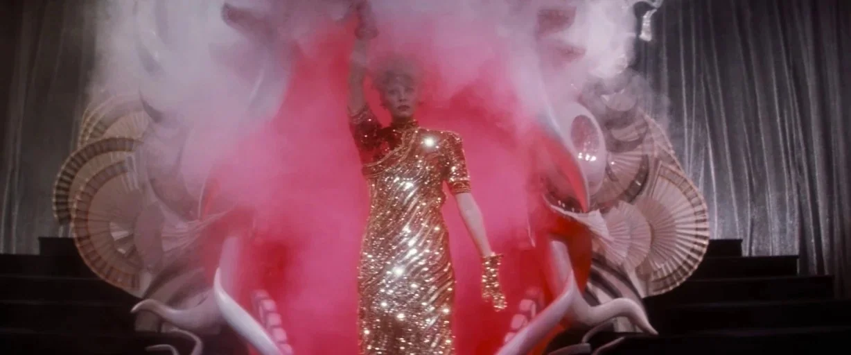 Indiana Jones and the Last Crusade (1989) movie still: medium — A woman stands in a bright red opening surrounded by large stylized fan props. The…