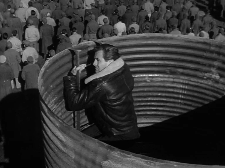 Stalag 17 (1953) movie still: medium — A man hides inside a large, corrugated metal pipe during a busy outdoor gathering. The surrounding area is…