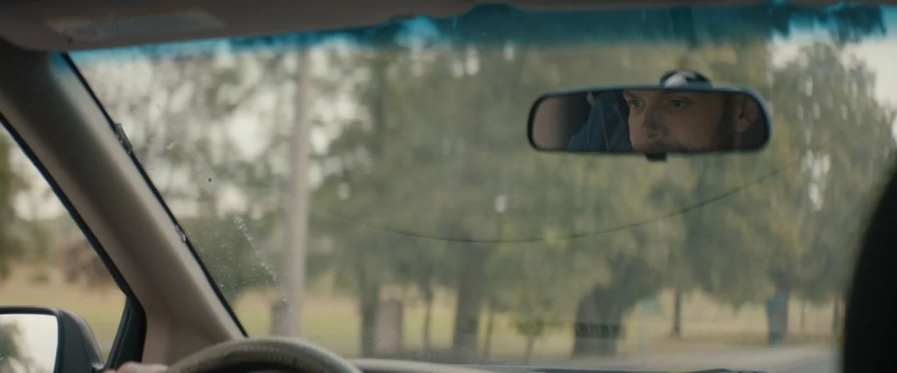Becky (2020) movie still: medium — The interior of a moving vehicle is viewed from the passenger or backseat perspective. A man is visible only…