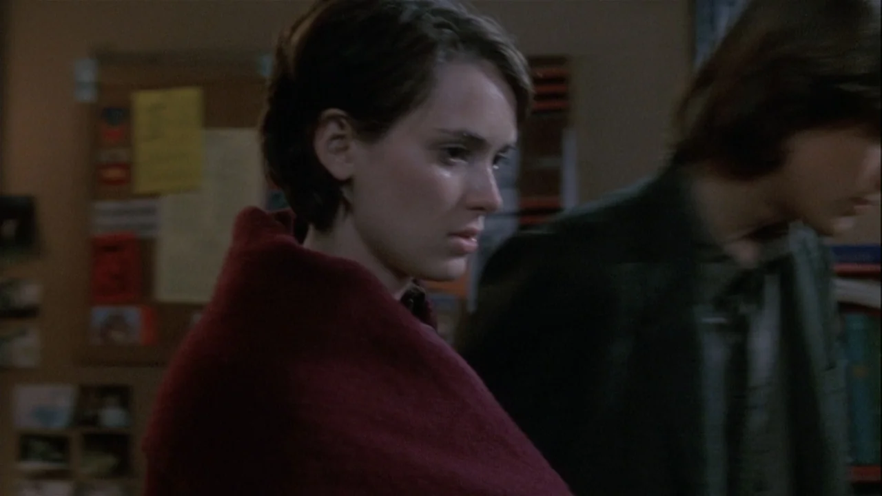 Boys (2021) movie still: medium — A dimly lit indoor space features a wall with a wooden bulletin board display. A woman in a dark red wrap stands…
