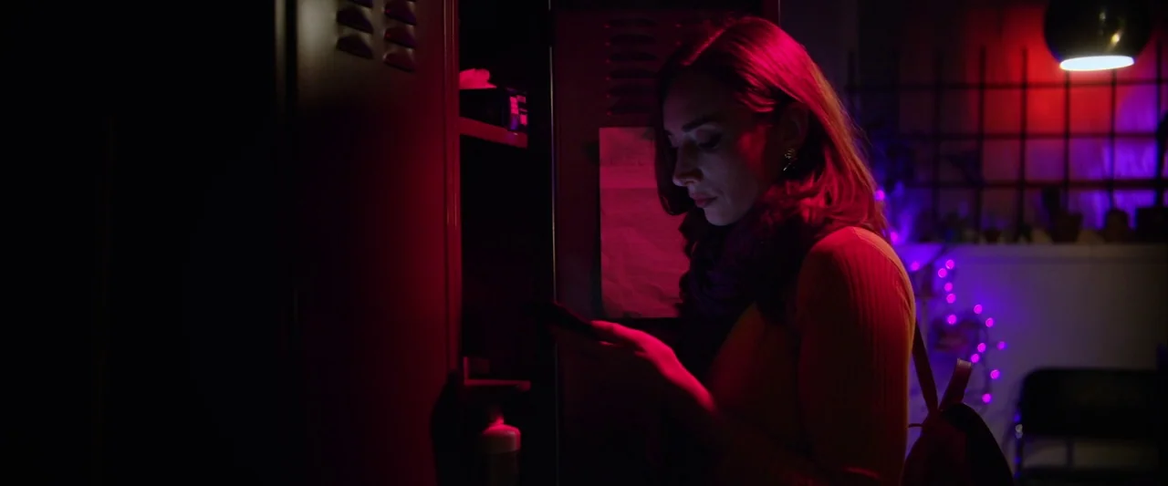 The Stylist (2020) movie still: medium — A dark space features a metal locker bathed in intense red and purple light. A young woman looks at her…