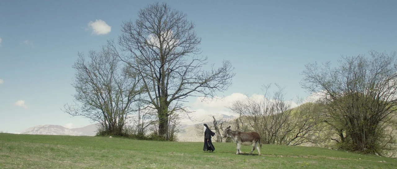 The Little Hours (2017) movie still: wide — An open grassy hillside is surrounded by bare deciduous trees and distant mountains. A nun wearing a…