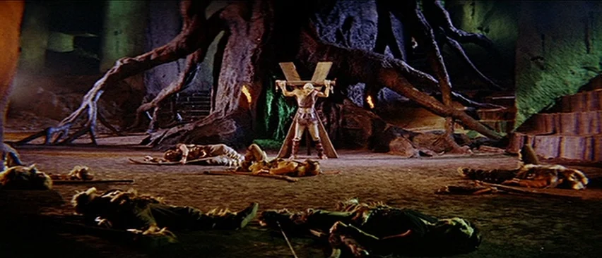 Erik The Conqueror (1961) movie still: wide — A subterranean cavern features a giant tree with exposed roots behind a wooden torture rack. Several…