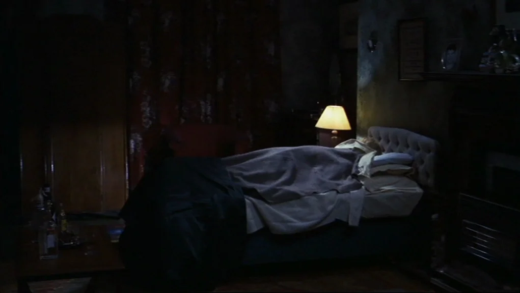 Frenzy (1972) movie still: medium — A dimly lit room with dark floral curtains and a bed frame. One person sits on the edge of the mattress near a…