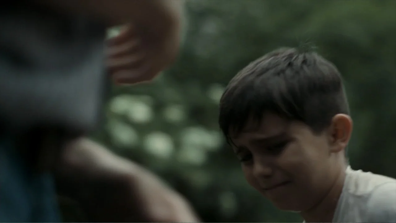 Alabama Snake (2020) movie still: closeup — A dense forest environment with soft natural light filtering through the canopy. A young boy stands in…