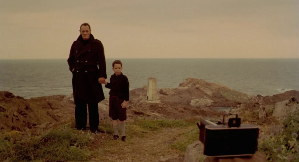 In a Glass Cage (1986) movie still: fullbody — A man and a young child stand together on a barren, rocky coastal cliff. The ocean and a distant…