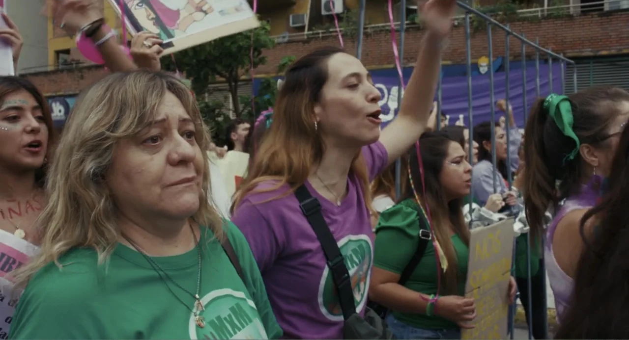 Belén (2025) movie still: medium — A group of women in green and purple t-shirts march down a city street. Several individuals are shouting and…
