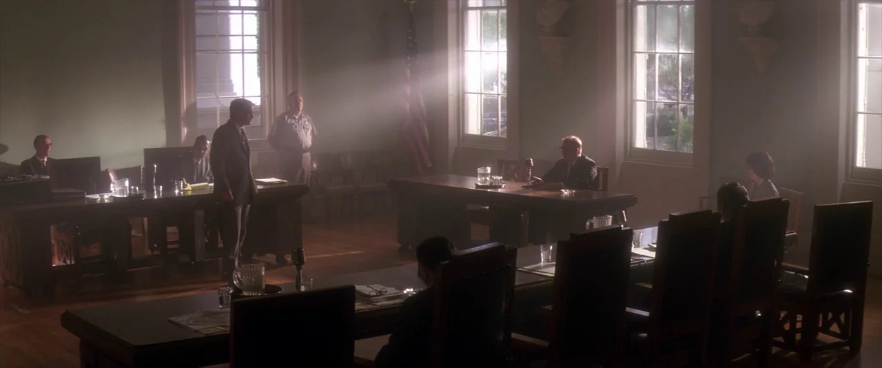 JFK (1991) movie still: wide — A dark, wood-paneled room features multiple long tables occupied by men in professional attire. Bright light pours…