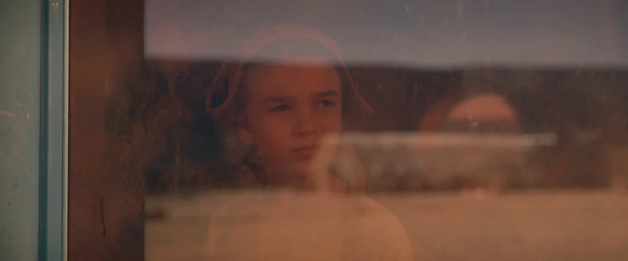 Settlers (2021) movie still: closeup — A young boy is visible through a dirty glass window pane. The reflection of the outdoors overlays the boy's…